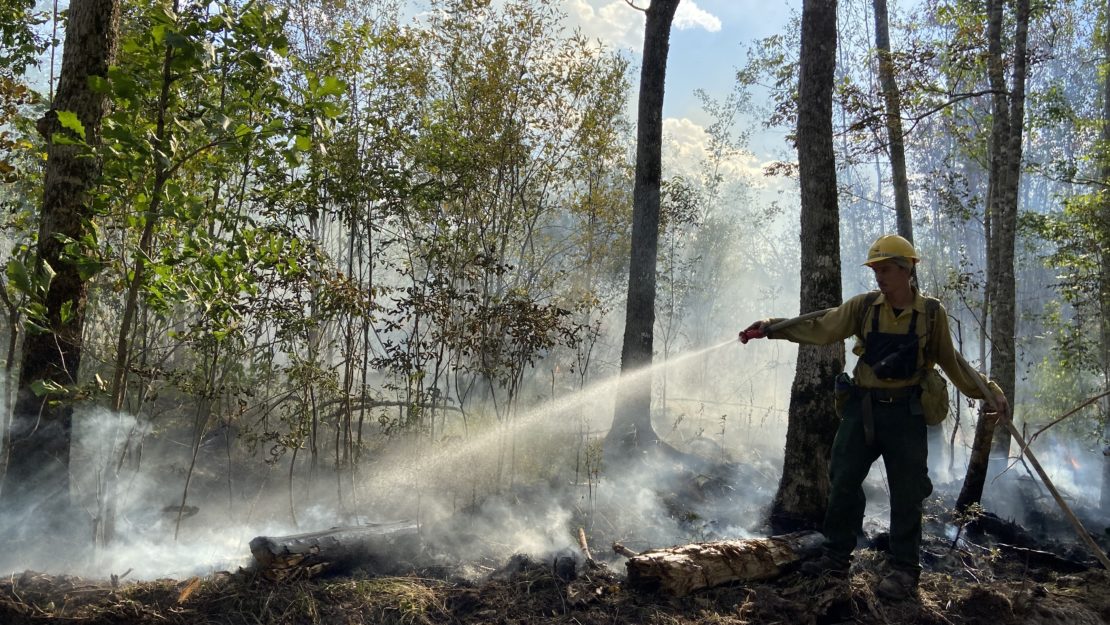 backlit firefighter using hose on smoldering fire.