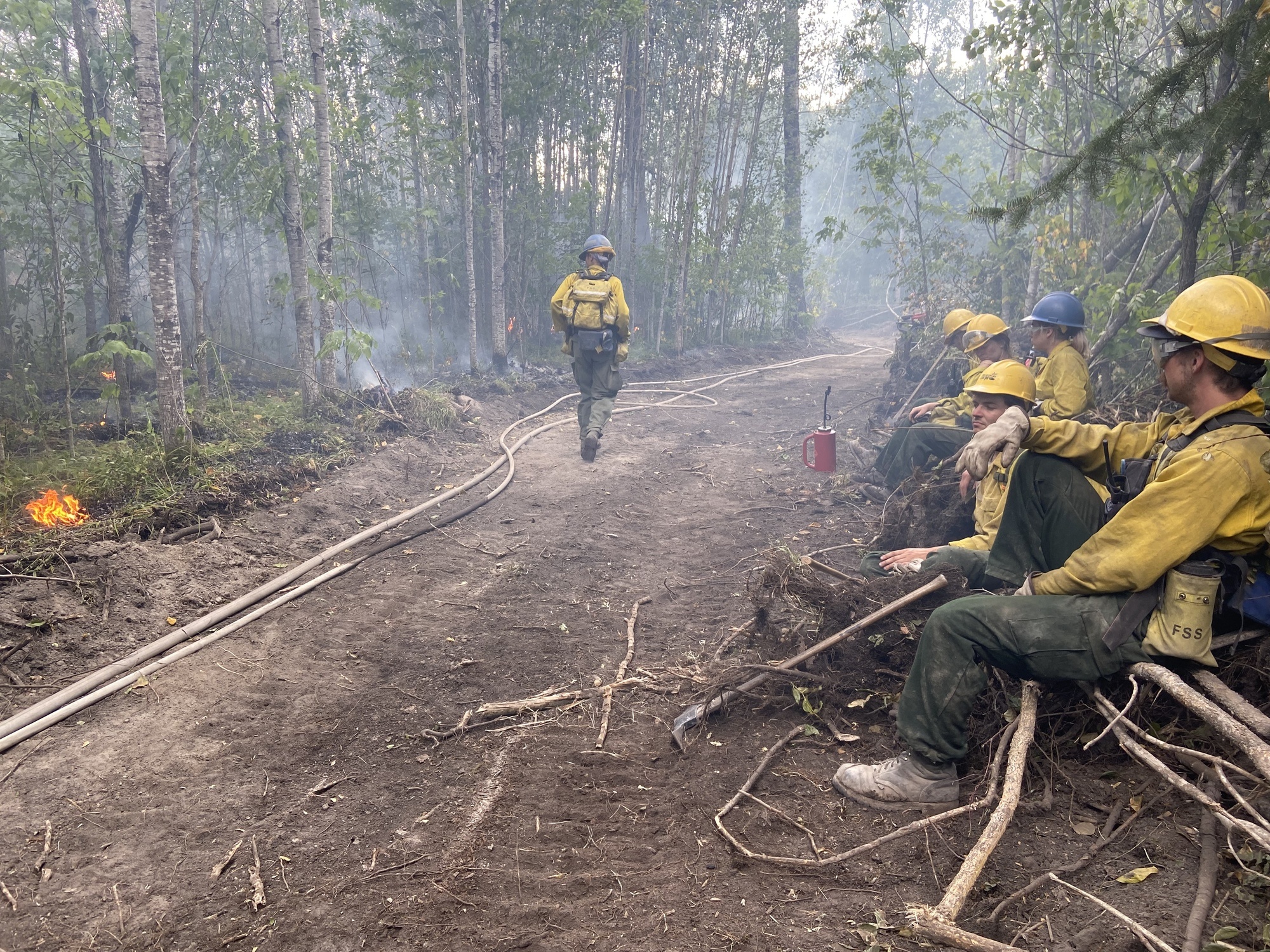 firefighters sitting on a dirt path with fire on the other side