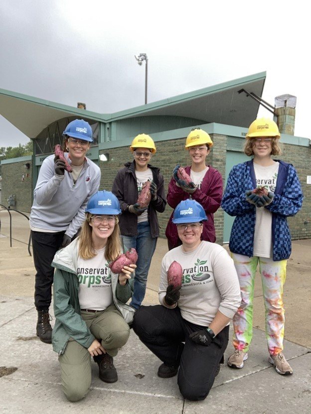 group of six youth posing for photo.Four individuals in a back line and two kneeling up front. Posing in front of a green park building. All holding a sweet potato and wearing hard hats and glasses