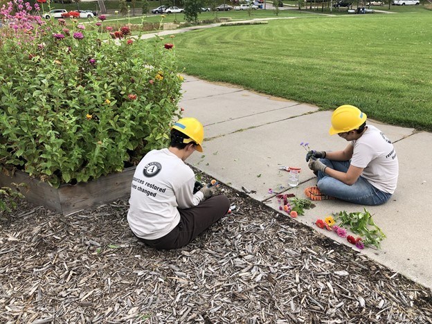 Two youth sitting in the ground arranging their flowers to put in the jars. Flower box next to youth and grassy field in the background.