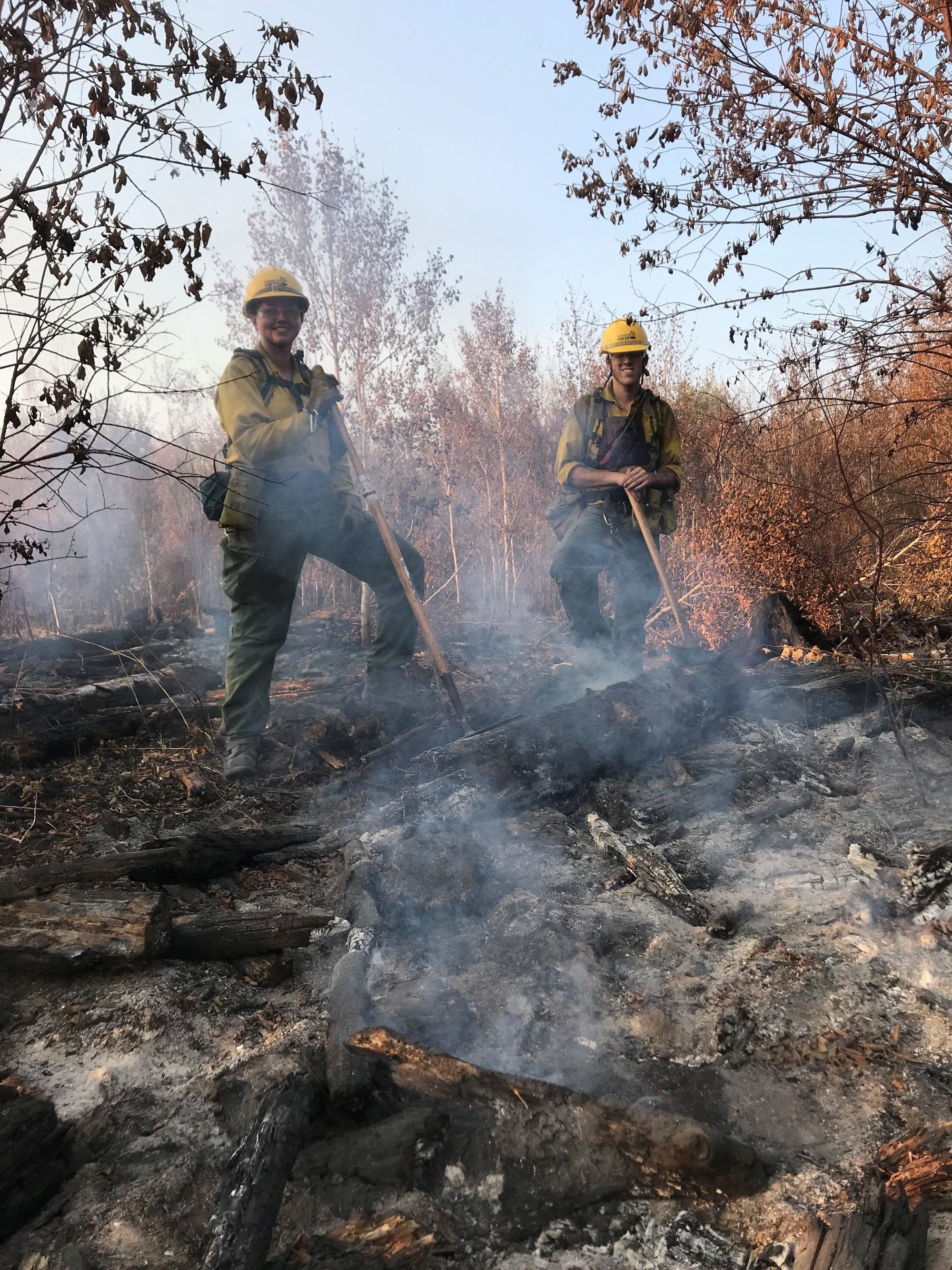 two firefighters standing in smoky blackened area holding hand tools