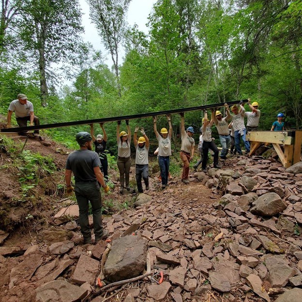 Team holding heavy beam above heads to span a streambed