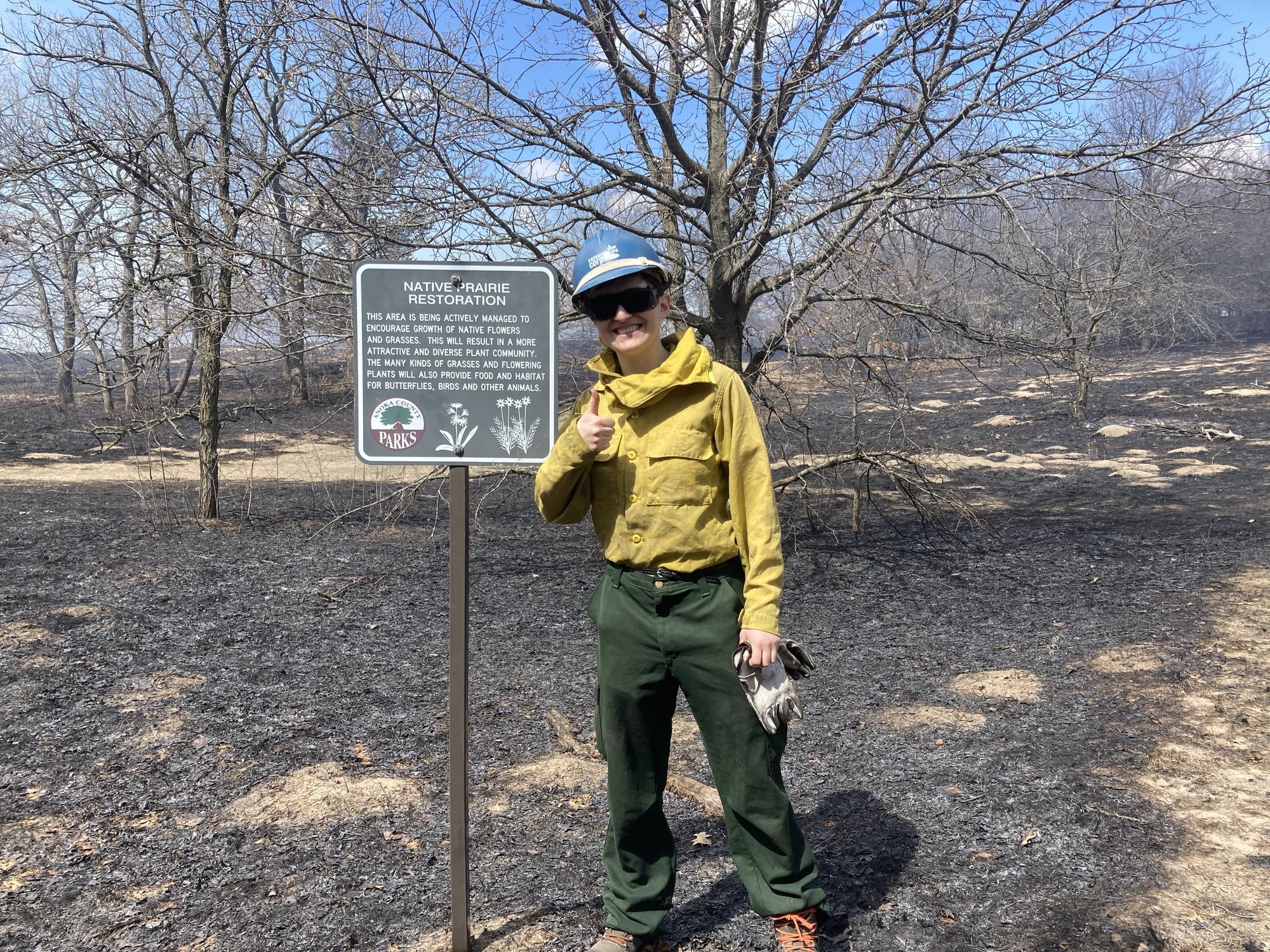 member in fire gear standing in front of blackened ground giving a thumbs up.