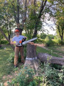member holding chainsaw by downed tree smiling.