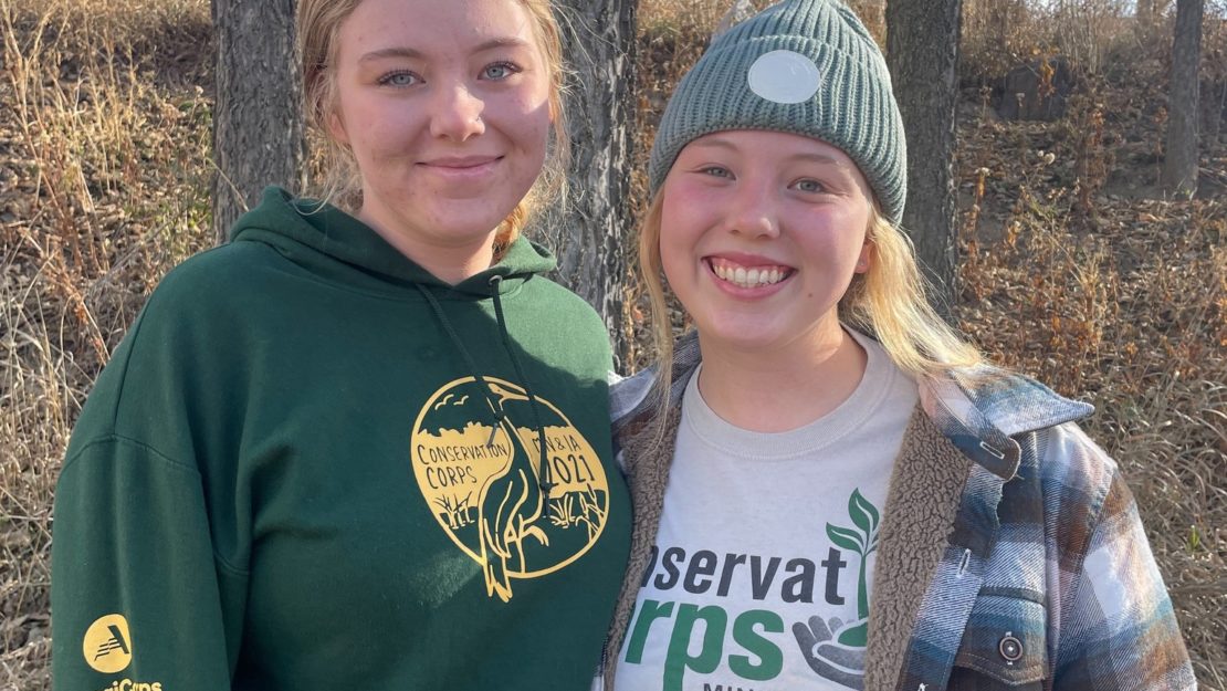 Two young people in conservation corps shirts in a forests standing side by side