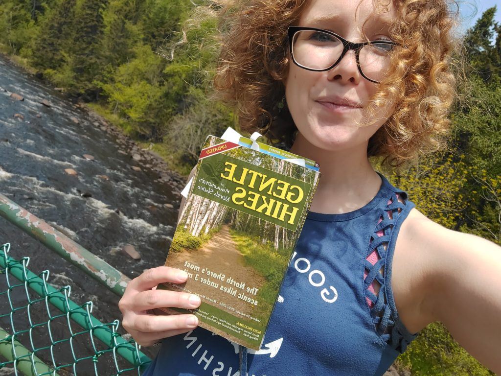 Someone holding up a book titled "Gentle Hikes" while standing in front of a river.