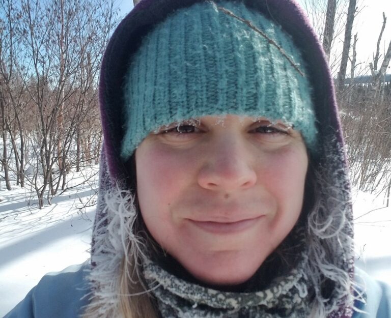 A woman in winter gear smiling at the camera in a snowy forest.