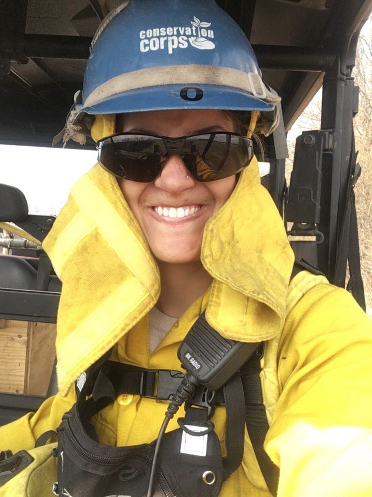 a person in firefighting gear with the Corps logo on their helmet smiles from the seat of an ATV.