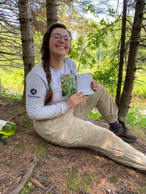 A person in a Corps shirt sits on the ground holding up a field guide.