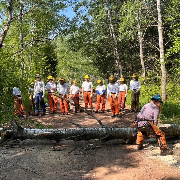Group of people in Corps gear watching someone how to use a chainsaw.