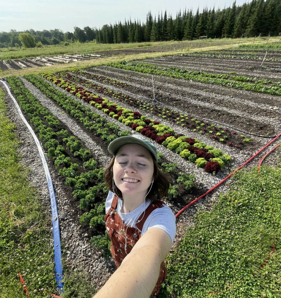 A Person taking a selfie smiling with their eyes closed standing in front of the lettuce crops.