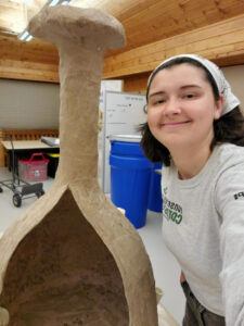 A person in a Corps shirt smiling beside a paper mache structure.