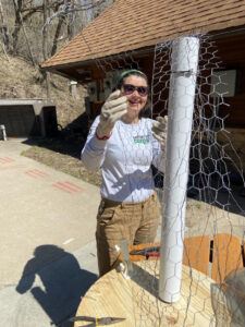 A person in sunglasses and a CCMI shirt wraps chicken wire fencing around the structure outside.
