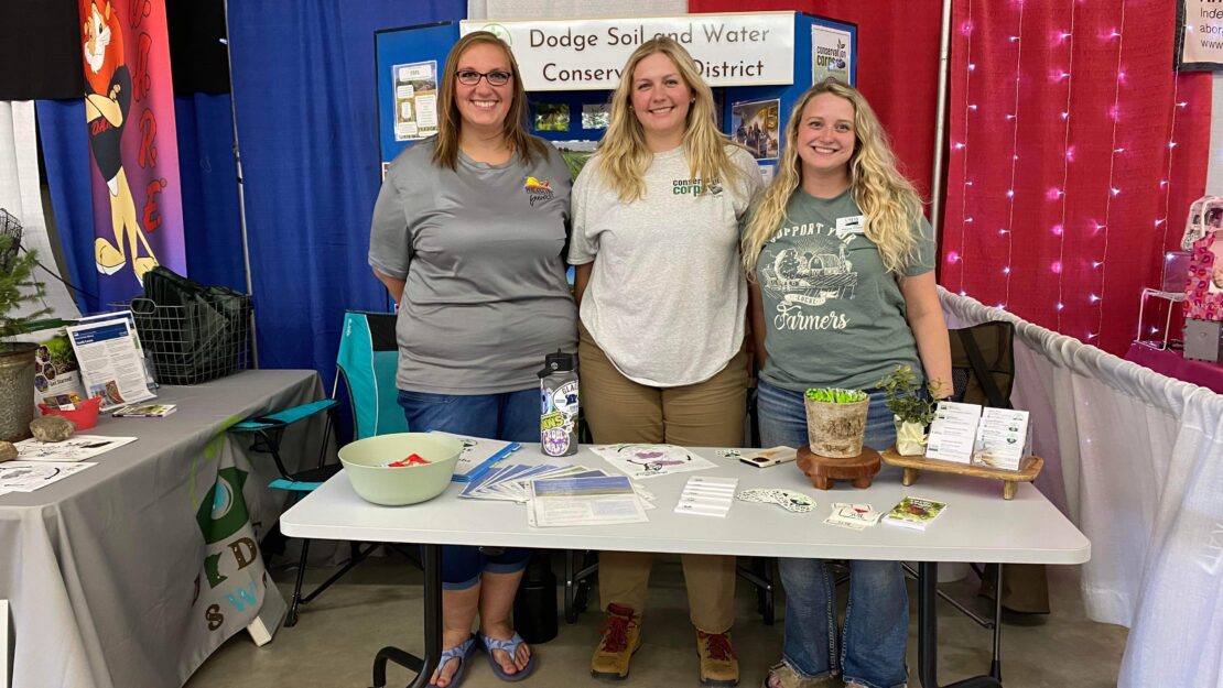 Three people, one in a Corps shirt, behind a table at a booth.