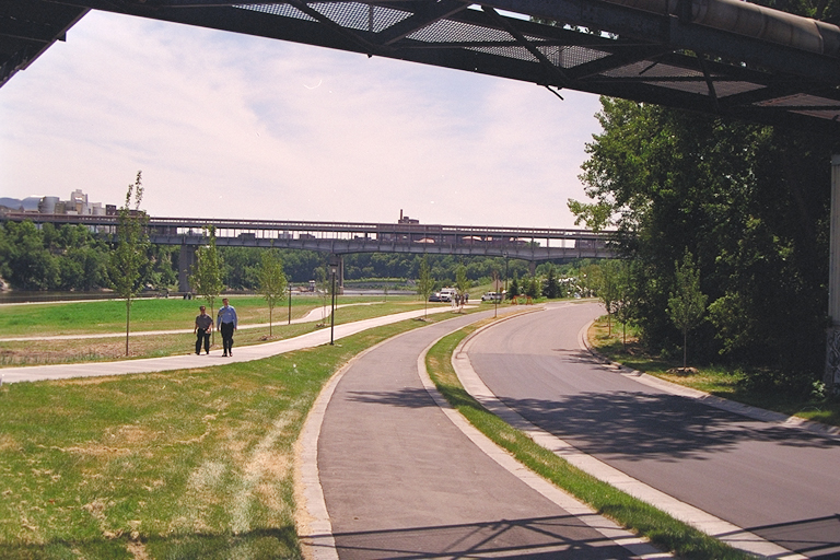 A green space in a river valley with two people walking along a path, a road runs alongside.