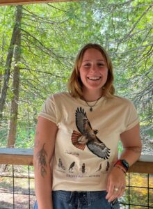 A person leaning against a fence in a forest wearing a bird shirt.