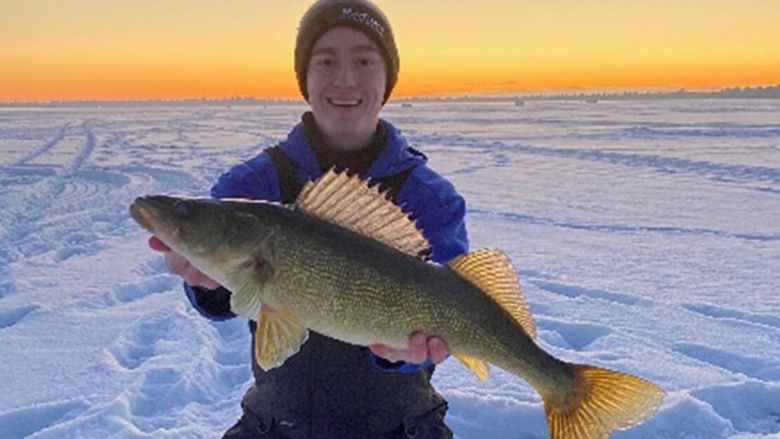 A person in winter gear kneeling on the ice holding a large fish.