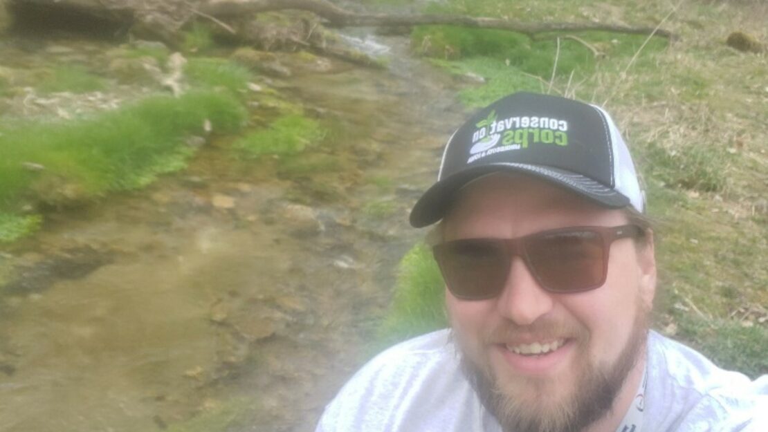 A person in a Corps hat by a stream