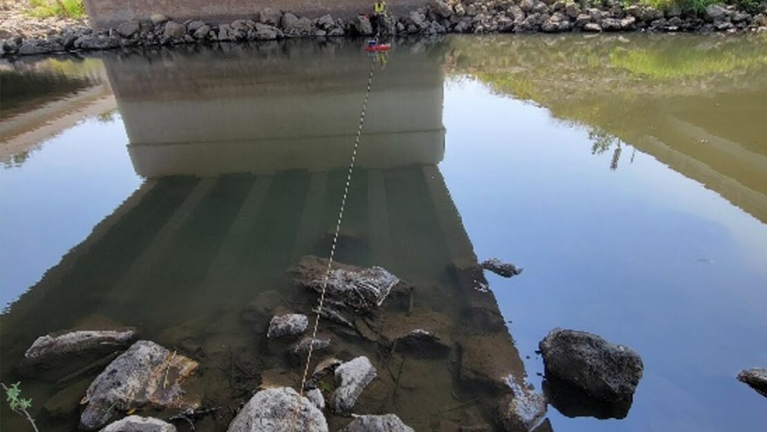 A person holding a string with another person holding the other end across a river. Both stand underneath a bridge over the river.