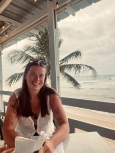 A person smiling with the ocean and a palm tree in the background.