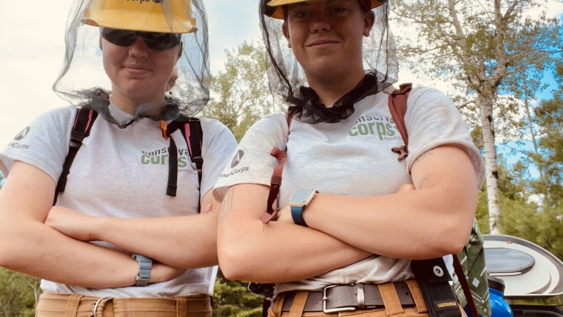 Two people in hard hats look at the camera with crossed arms