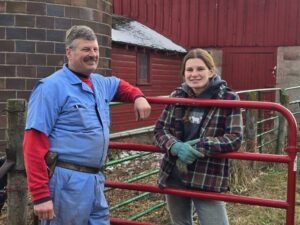 Two people standing by a red gate with farm buildings in the background